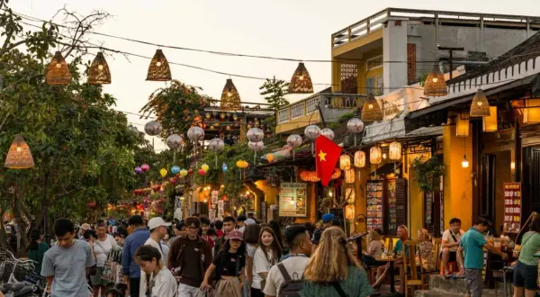 Bunte Lichter und Menschenmassen beleuchten die **Hoi An Altstadt**. Ein vietnamesisches Banner weht über einer belebten Straße voller Geschäfte und Restaurants.