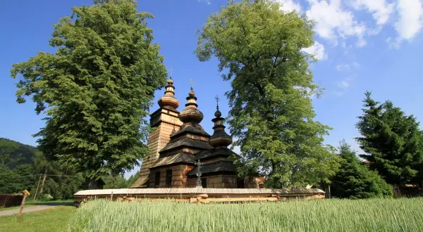 Drewniana cerkiew z kopułami na tle zieleni i błękitnego nieba, otoczona polami zbóż. Widok na Beskid Niski.