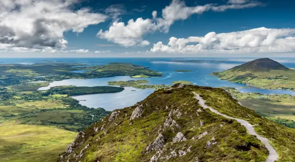 Vue panoramique du Connemara depuis Galway. Un sentier serpente sur une colline verdoyante, menant à des lacs bleus et des îles sous un ciel nuageux.