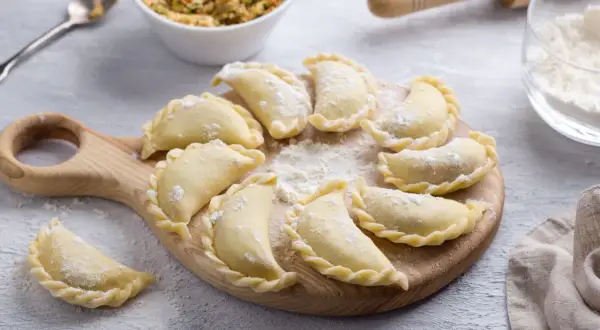 Gotowe do gotowania pierogi ruskie ułożone na desce, posypane mąką. Obok miska z farszem i wałek.