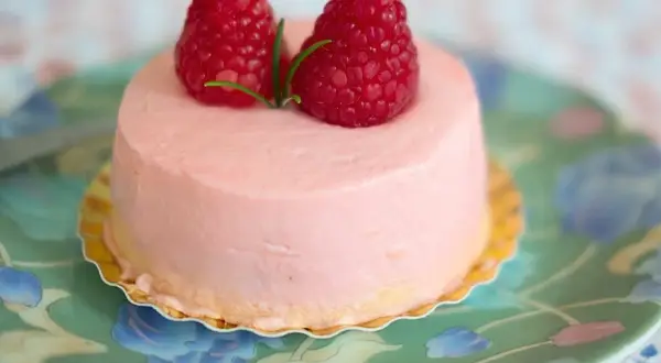 Delikatny krem malinowy z mascarpone do tortu, ozdobiony dwoma świeżymi malinami i gałązką rozmarynu, na zielonym talerzyku w kwiaty.