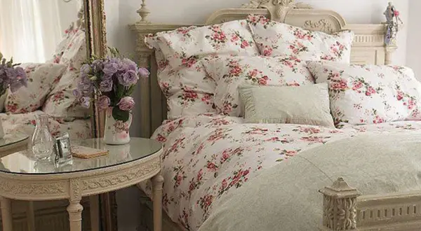 En säng med blommig shabby chic inredning, prydd med rosor och lavendel. Ett runt spegelbord med en vas full av blommor.