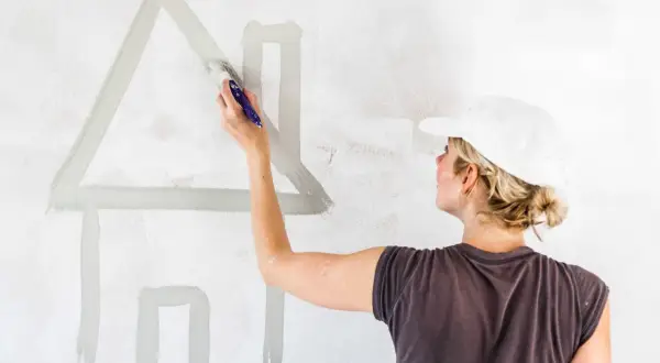 Donna con cappellino bianco dipinge una casa stilizzata su muro. Calcolare quanti litri di pittura per 50 mq.
