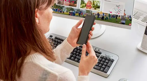 Kobieta trzyma telefon, szukając informacji, jak podłączyć telefon do monitora. Na ekranie komputera widać zdjęcia.