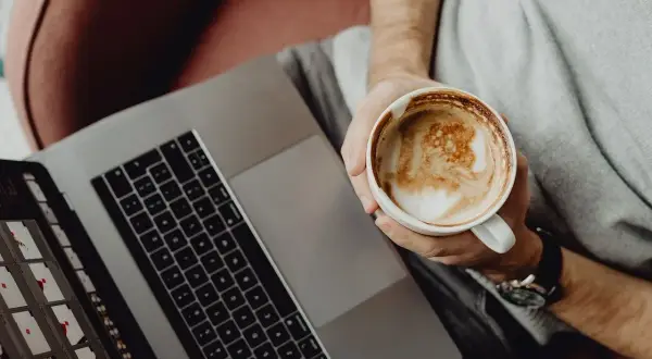 Osoba z kawą na laptopie, ucząca się Mac OS dla początkujących.