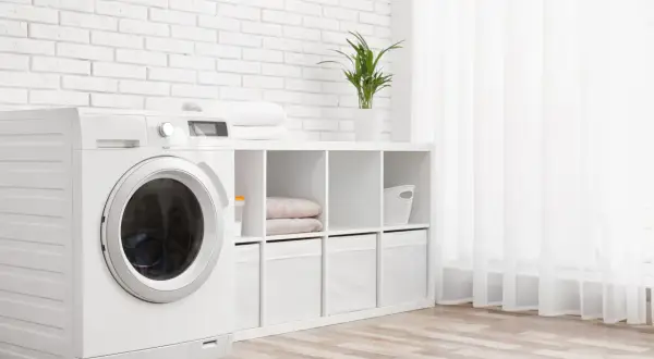 Weiße Waschmaschine mit beladener Trommel steht neben einem Regal mit Handtüchern und Aufbewahrungsboxen. Die Waschmaschine wandert nicht, sie steht fest.