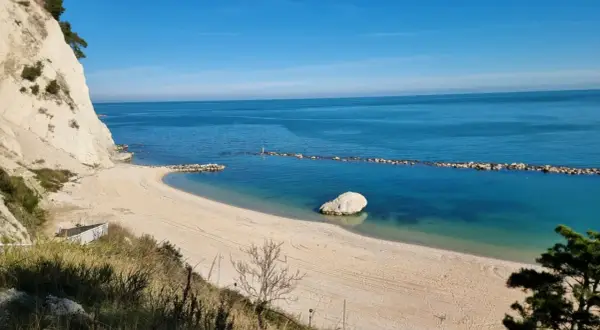 Spiaggia dorata e mare cristallino sulla Riviera del Conero, con una scogliera bianca a picco sull'acqua e un grande masso solitario.