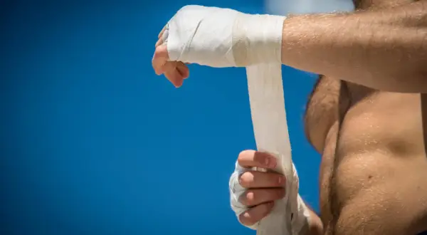 A boxer wraps his hands, preparing for a fight under a bright blue sky.