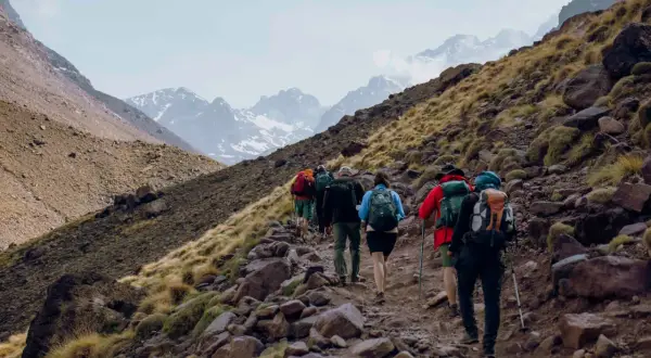 Grupa turystów wspina się kamienistym szlakiem na zboczu gór w Maroku. W tle widać ośnieżone szczyty, prawdopodobnie Jebel Toubkal.
