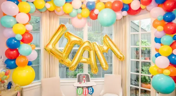 Złote balony na roczek tworzą napis "LIAM" na tle kolorowej girlandy z balonów.