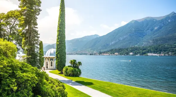 Giardino lussureggiante con gazebo bianco e cipressi slanciati, affacciato sul blu intenso del lago di Como, con montagne verdi sullo sfondo.