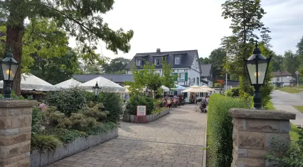 Gemütlicher Biergarten mit Sonnenschirmen und Gästen. Ein Schild weist auf den "Kleine Schweiz Velbert Rundweg" hin.