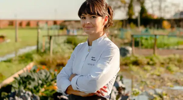 Chiara Pavan, chef con grembiule e giacca bianca, sorride con le braccia conserte in un orto.