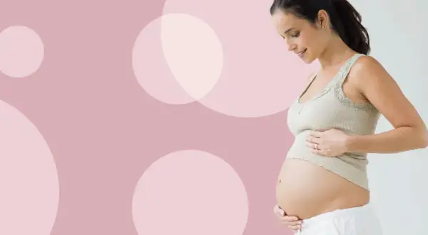 Uśmiechnięta kobieta w ciąży dotyka brzucha. Waga dziecka w 27 tygodniu ciąży jest już znacząca.