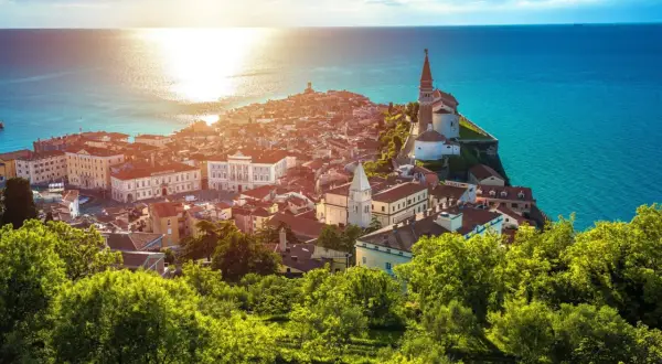 Sonnenuntergang über der malerischen Piran Altstadt, mit ihren roten Dächern und der Kirche, die auf einer Landzunge ins türkisfarbene Meer ragt.