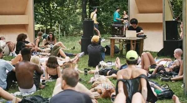 Un gruppo di persone rilassate sull'erba, ascoltando musica a un butik festival. Un DJ mixa con un laptop, circondato da altoparlanti e una struttura in legno.