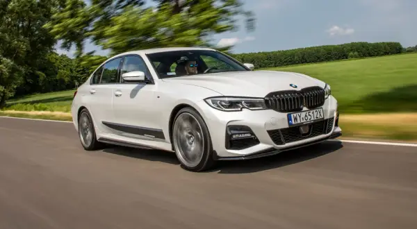 Białe BMW 320d xDrive pędzi po drodze, otoczone zielenią pól i drzew.