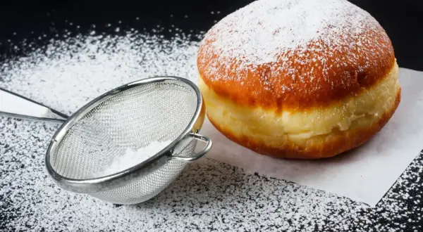 Puszysty pączek posypany cukrem pudrem, obok sitko z cukrem. Idealne na tłusty czwartek, pyszne pączki!