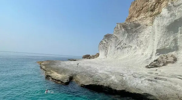 Dos personas nadan en las **calas agua amarga**, con acantilados blancos y mar turquesa bajo un cielo azul.