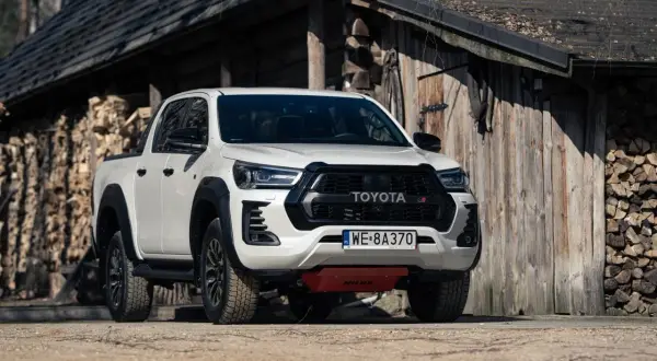 Biały Toyota Hilux GR Sport z czarnymi dodatkami, zaparkowany przed drewnianym budynkiem z polanami drewna.