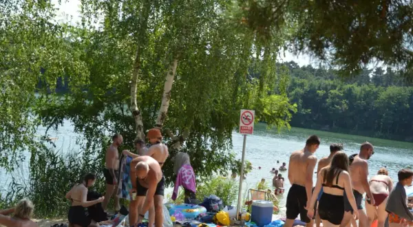 Ludzie odpoczywają nad jeziorem, niektórzy kąpią się, inni siedzą na plaży. W tle widać las. To idealne miejsce na relaks, jakby czas się zatrzymał, niczym w starożytnym rydwanie.