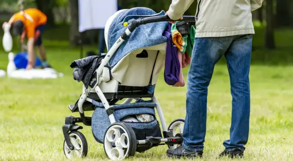 Osoba na urlopie rodzicielskim pcha wózek dziecięcy po trawie. W tle widać bawiących się ludzi.