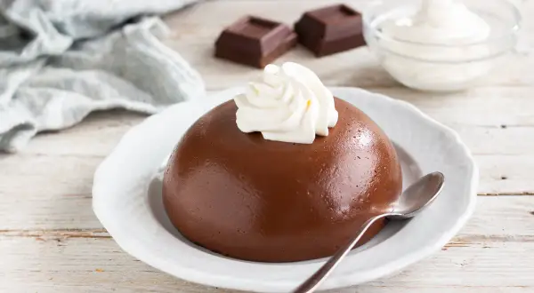 Budino al cacao liscio e lucido, guarnito con panna montata, su un piatto bianco. Accanto, cubetti di cioccolato e una ciotola di panna.
