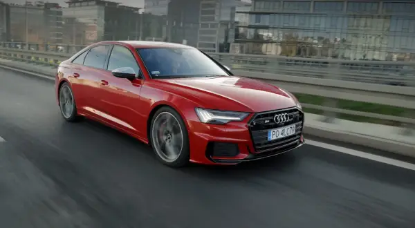 Czerwone, miękka hybryda Audi S6 sunie po drodze, za kierownicą siedzi kierowca w okularach.