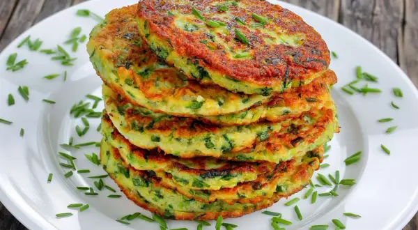 Pile de galettes de courgette dorées, parsemées de ciboulette fraîche sur une assiette blanche. Un délice végétal.