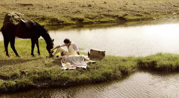 Pareja en un picnic junto a un río, con un caballo pastando. Un momento idílico para disfrutar de rutas en caballo y la naturaleza.