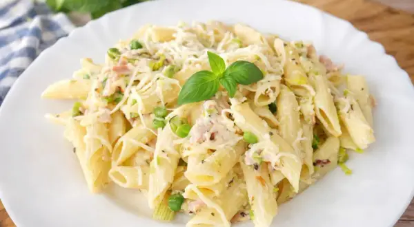Penne z kremowym sosem, kawałkami szynki, groszkiem i startym parmezanem, udekorowane listkiem bazylii. Pyszny makaron z szynką.