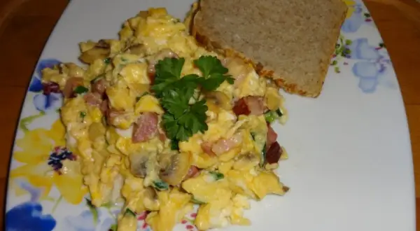 Pyszna jajecznica z pieczarkami, boczkiem i szczypiorkiem, udekorowana natką pietruszki, podana z kromką chleba.
