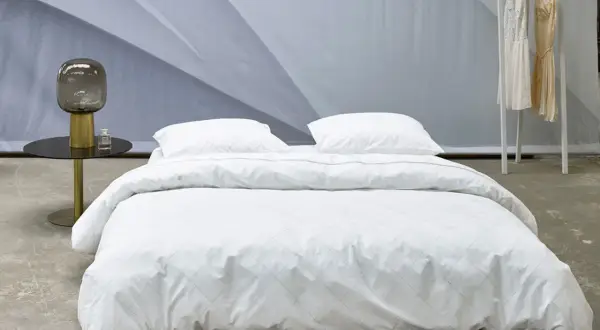 Ein Bett mit weißer Perkal-Bettwäsche, gesteppt mit feinen Linien. Daneben ein Nachttisch mit Lampe und Parfüm.