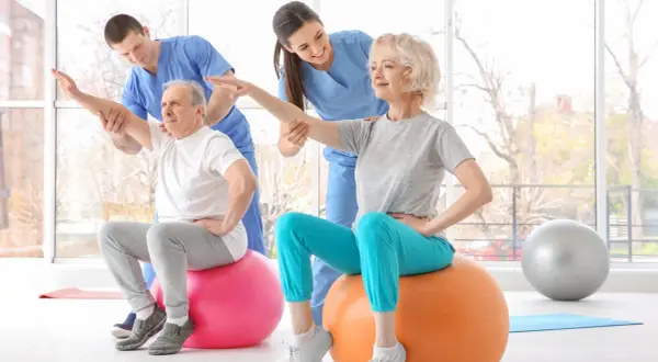 Rehabilitacja dla seniorów: starsza pani i pan ćwiczą z fizjoterapeutami na piłkach gimnastycznych, poprawiając kondycję.