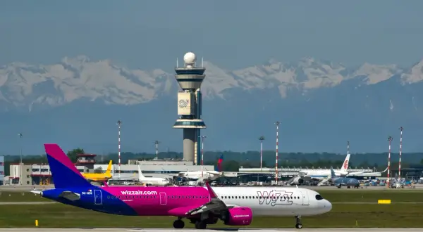 Samolot Wizz Air na płycie lotniska z wieżą kontroli lotów i górami w tle.