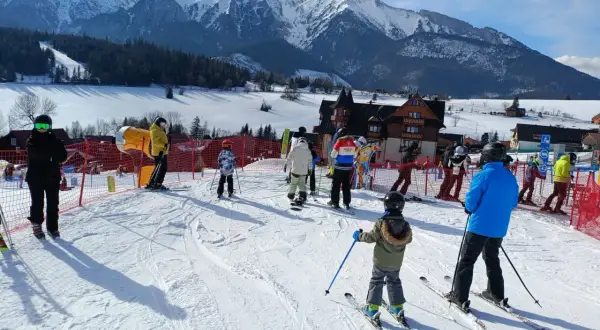 Ludzie na nartach i snowboardach w słoneczny dzień w ski centrum Strachan, z majestatycznymi ośnieżonymi górami w tle.