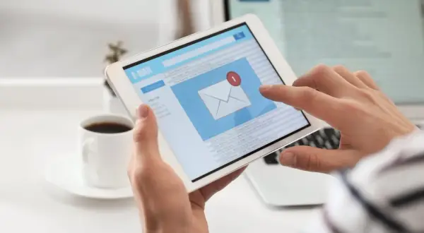 Ręce trzymają tablet z ekranem poczty e-mail, pokazującym jak napisać maila po angielsku. Obok kawa.