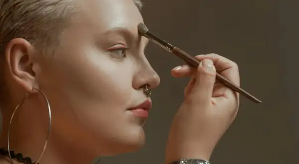 Kobieta z krótkimi blond włosami i kolczykiem w nosie nakłada bronzer na czoło, ucząc się, jak wyszczuplić twarz bronzerem.
