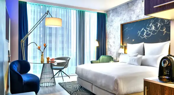 Nowoczesny pokój w hotelu NYX Warszawa: łóżko z białym pościelą, stolik kawowy, granatowe fotele i zielona sofa.
