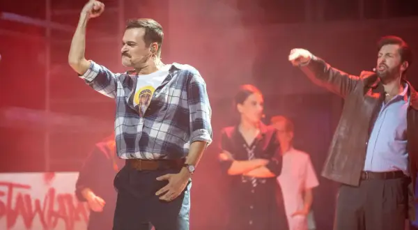 Aktorzy na scenie musicalu, jeden z podniesioną pięścią, w tle inne postacie i napisy.