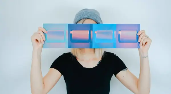 Osoba w czapce i czarnej koszulce trzyma przed twarzą panel z czterema prostokątami w odcieniach niebieskiego i różowego. Inspiracja do studiów artystycznych kierunki.