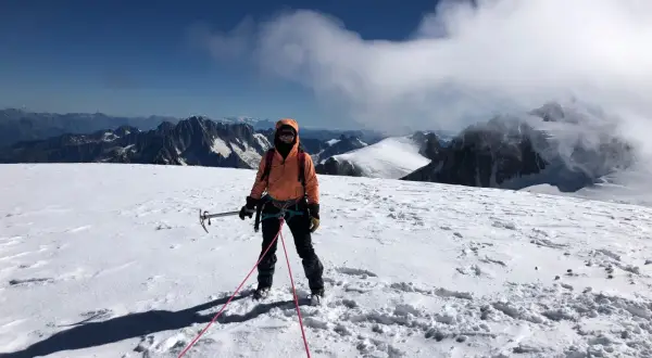 Une alpiniste, équipée pour l'ascension d'une montagne, se tient sur un sommet enneigé, le regard tourné vers l'horizon.