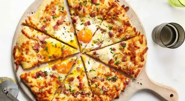 Kawałki pizzy carbonara z jajkiem sadzonym, boczkiem i szczypiorkiem, ułożone na desce.