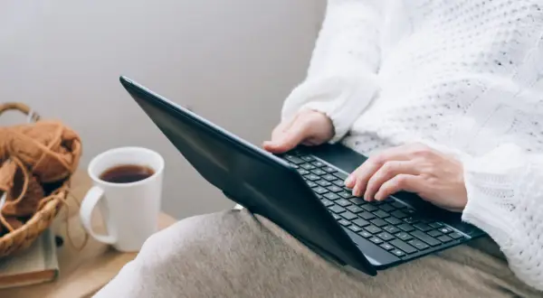 Osoba w białym swetrze pisze na laptopie, obok kubek z kawą i koszyk z włóczką.