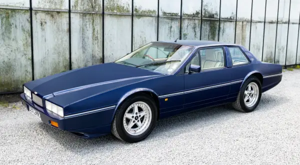 Granatowy Aston Martin Lagonda z lat 80. na tle industrialnej ściany.