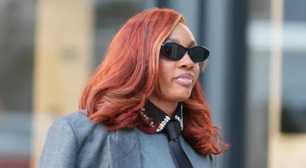 Frau mit feuriger, **cooler Haarfarbe** und stylischer Sonnenbrille trägt einen grauen Blazer mit Krawatte und glitzerndem Kragen.
