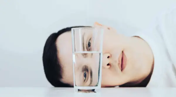 Un rostro se refleja en un vaso de agua, mostrando una dualidad entre el pesimista y el optimista.
