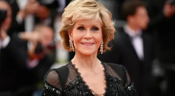 Uśmiechnięta Jane Fonda przed operacjami, w czarnej, ozdobnej sukni i długich kolczykach.