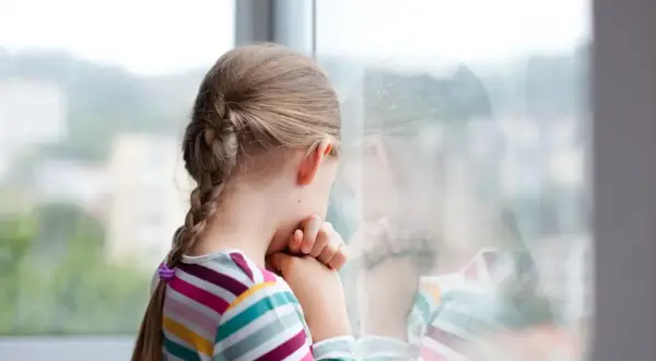 Smutna dziewczynka z warkoczem patrzy przez okno. Czuje się samotna, bo dzieci nie chcą się bawić z moim dzieckiem w szkole.