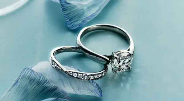 Dos anillos de compromiso, uno con un gran diamante y otro con pequeñas gemas, descansan sobre pétalos de flor azul. Un anillo casado para siempre.